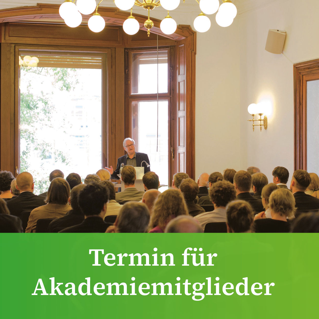 Sitzungssaal der Akademie mit großem Erkerfenster und Kronleuchter. Im Vordergrund sieht man Zuhörerinnen und Zuhörer, am Pult vorn spricht ein Redner. Unten im Bild steht vor grünem Hintergrund "Termin für Akademiemitglieder".