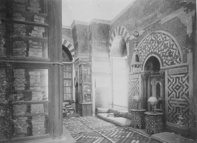 Schwarz-weiß-Foto einer alten Bibliothek mit verzierten Rundbögen, vollen Bücherregalen und Fußbodenmosaiken