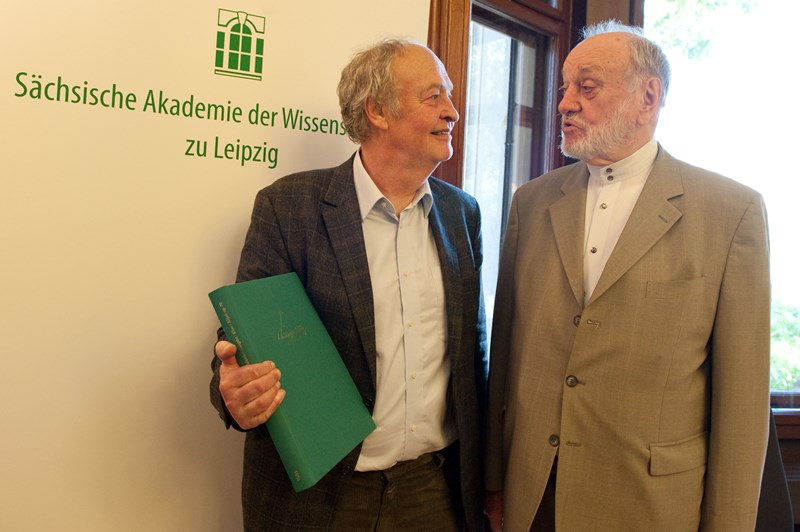 Zwei männliche Personen unterhalten sich, einer hält ein grünes Buch in den Händen. Im Hintergrund sieht man einen Aufsteller mit dem Logo der Sächsischen Akademie der Wissenschaften zu Leipzig.