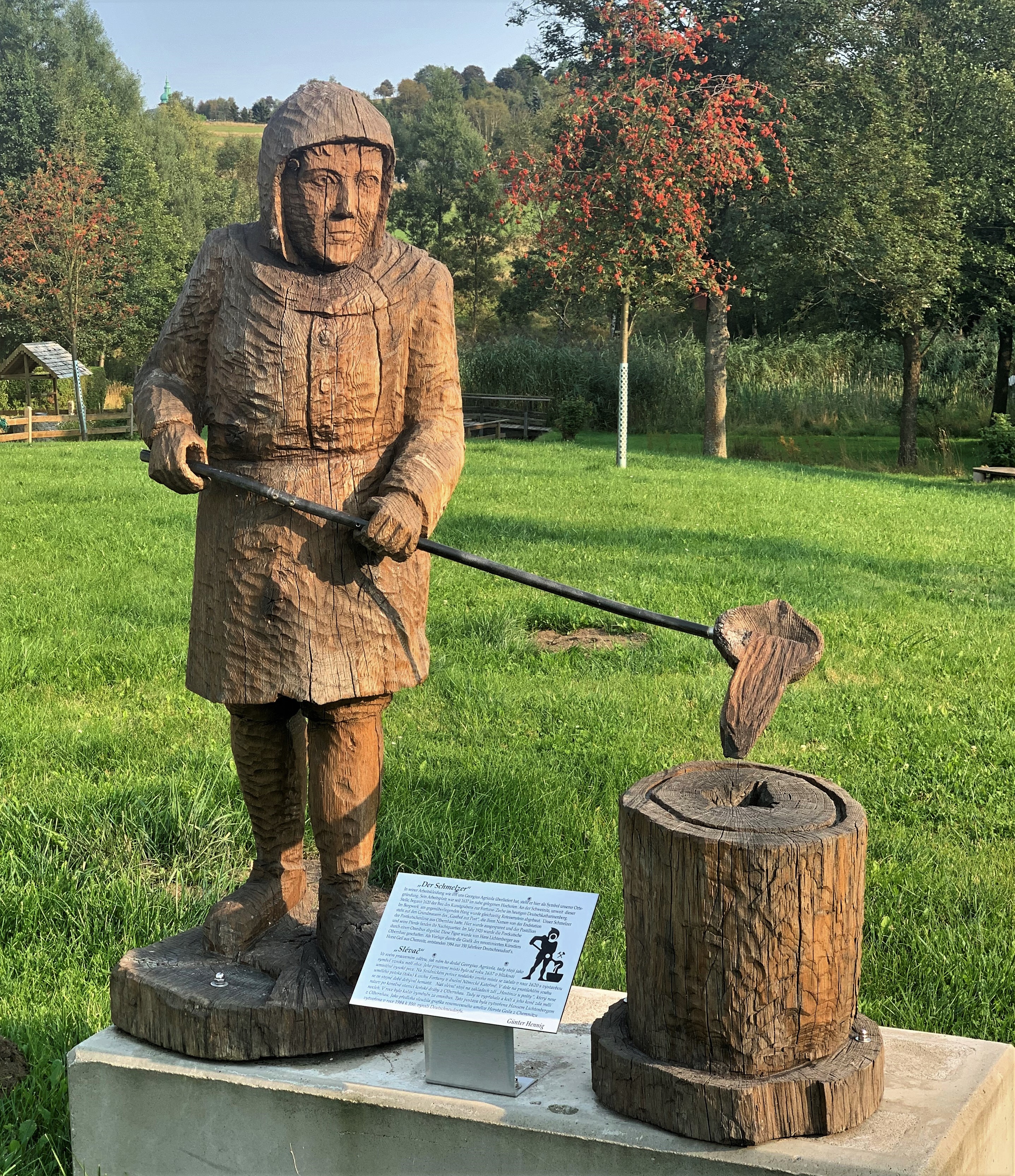 Wooden sculpture of a smelter at work. There is a meadow and there are trees in the background.